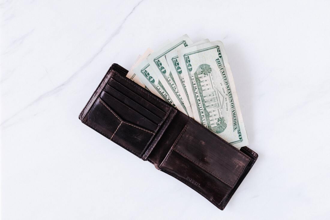 A dark colored wallet on a light background with a few $20 cash bills spilling out